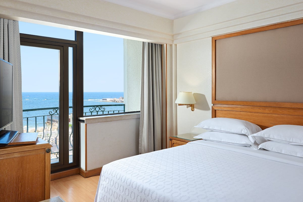 A hotel room with a large bed, a desk with a laptop, a nightstand with a lamp, and a view of the sea through a balcony.