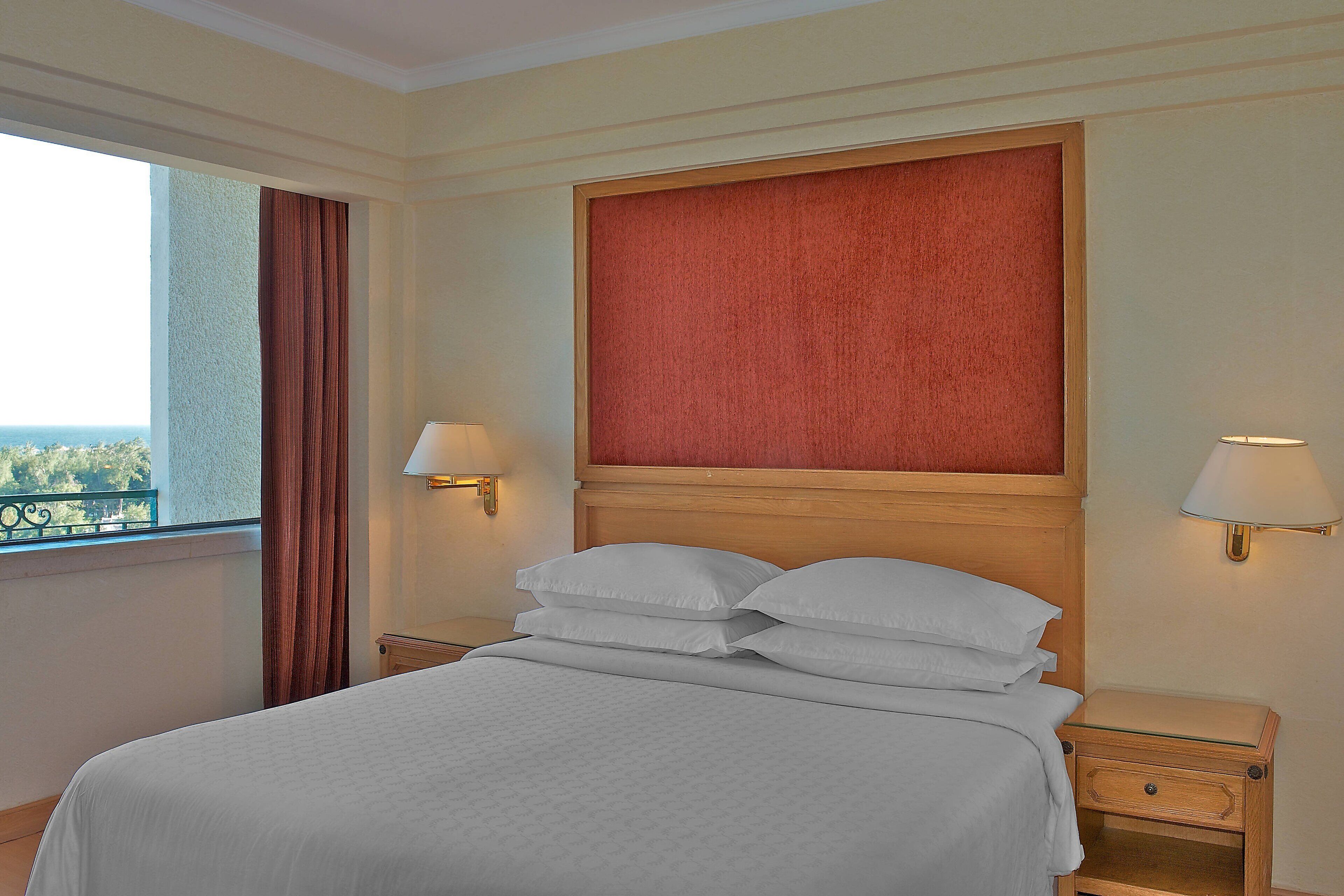 Quarto de hotel com uma cama grande, duas mesas de cabeceira com luminárias, cabeceira vermelha e vista para o exterior.