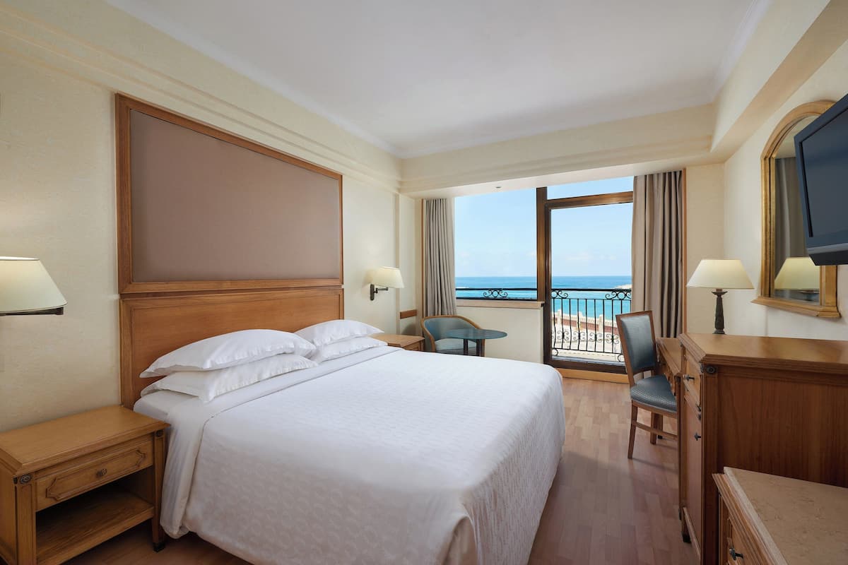 A hotel room with a large bed, a desk, a chair, a TV, and a balcony with a view of the sea.