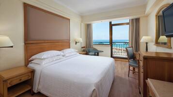 A hotel room with a large bed, a desk, a chair, a TV, and a balcony with a view of the sea.