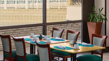 Un comedor con vista a una cerca blanca y edificios. Hay seis sillas de madera con cojines verdes y cojines blancos con lunares. La mesa está servida con tazas, platillos y una piña.
