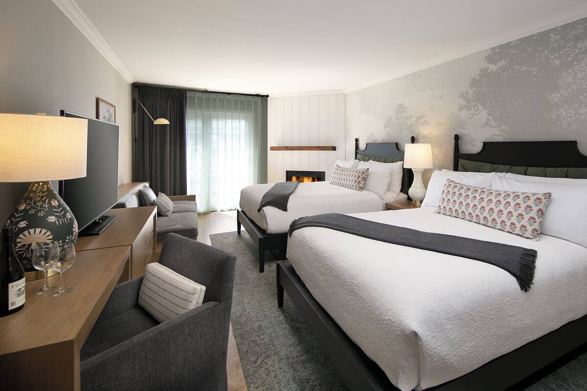 A hotel room with two beds, a desk, a chair, a lamp, and a fireplace.