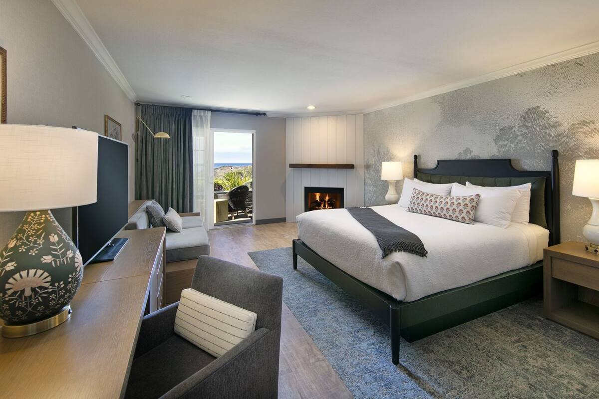 A modern hotel room with a large bed, two bedside lamps, a desk with a chair, a sofa, and a fireplace.