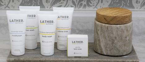 Un conjunto de artículos de tocador LATHER® de estilo moderno, incluyendo crema hidratante, champú, gel de ducha y jabón facial, sobre una superficie de piedra con un recipiente de madera.