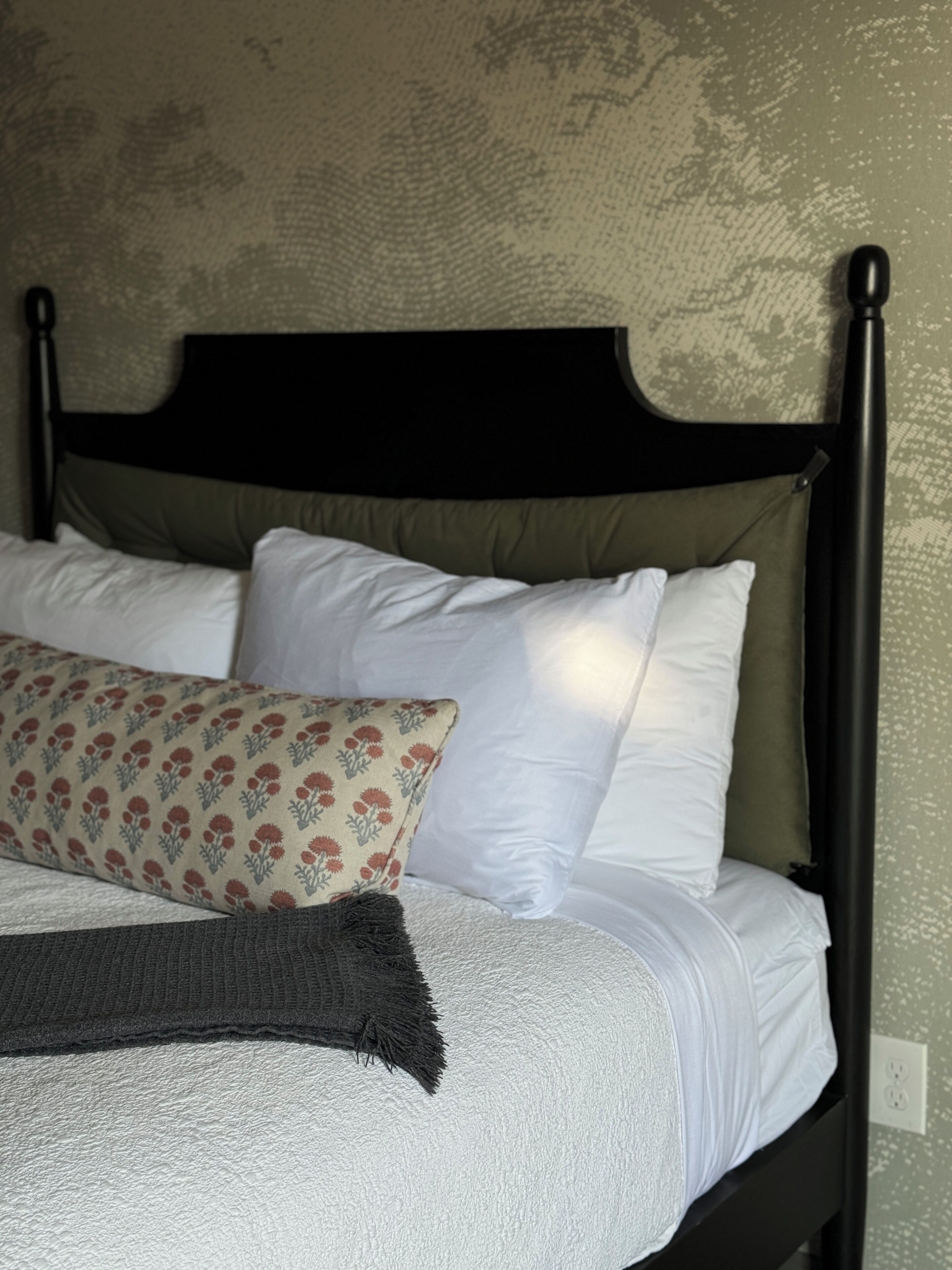 A bed with a dark headboard, white and patterned pillows, and a gray throw blanket.