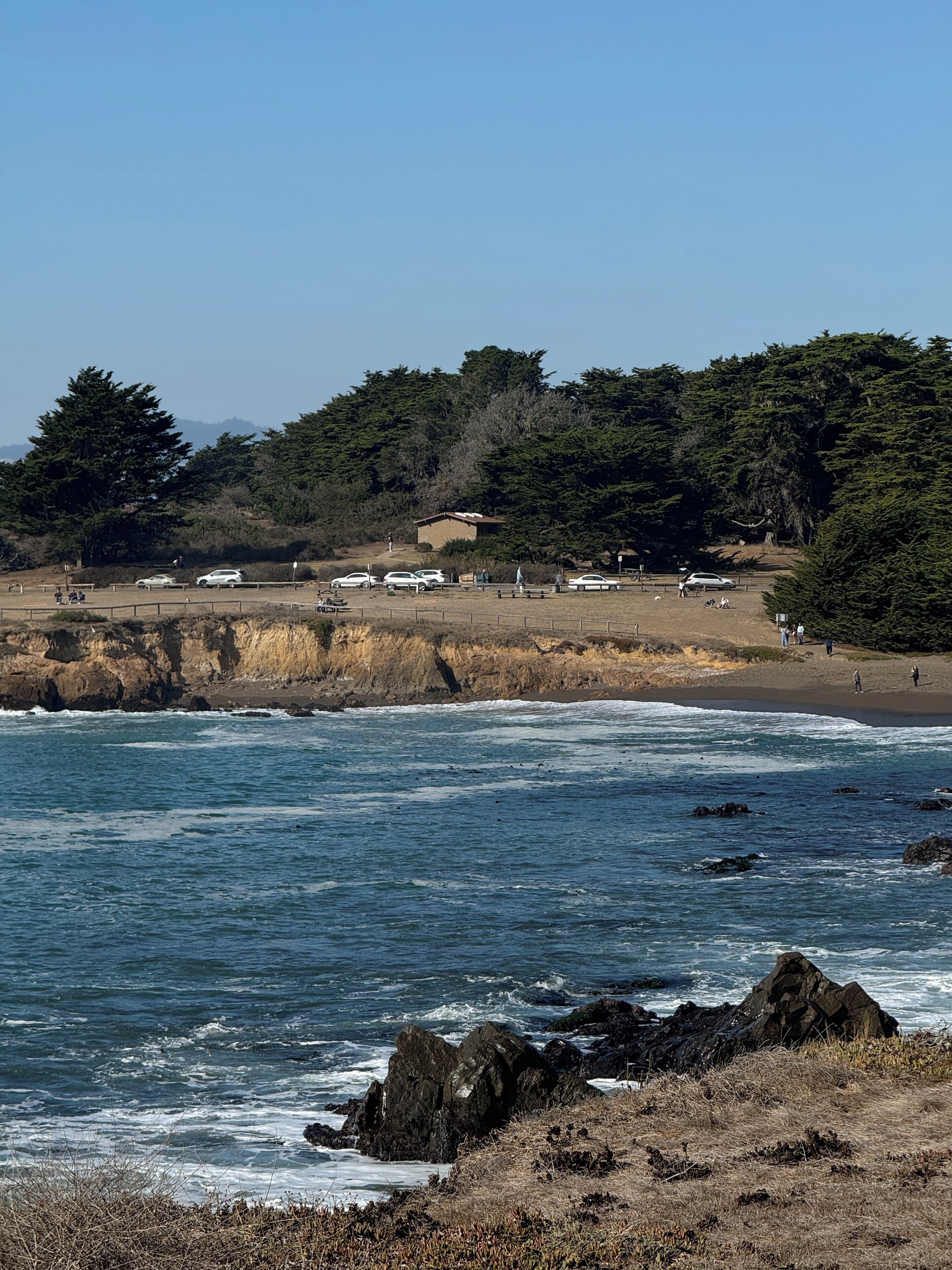 Une zone côtière avec un rivage rocheux, un parking où sont garées plusieurs voitures et une zone herbeuse où se trouvent des personnes.