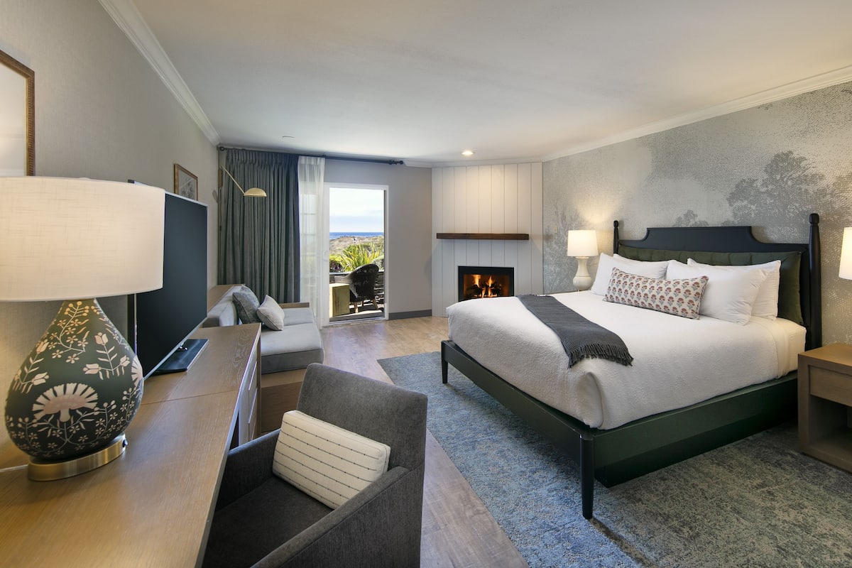 A modern hotel room with a large bed, two bedside tables with lamps, a sofa, a desk, and a fireplace.
