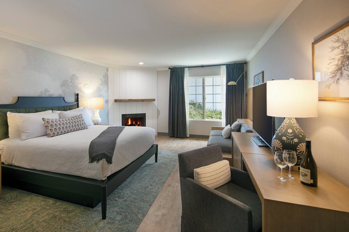 A modern hotel bedroom with a large bed, bedside lamps, a fireplace, a sofa, a desk with a lamp, and a view of the outdoors.