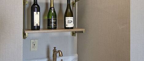 Un baño con lavamanos de mármol, grifo dorado y tres botellas de vino sobre una estantería.