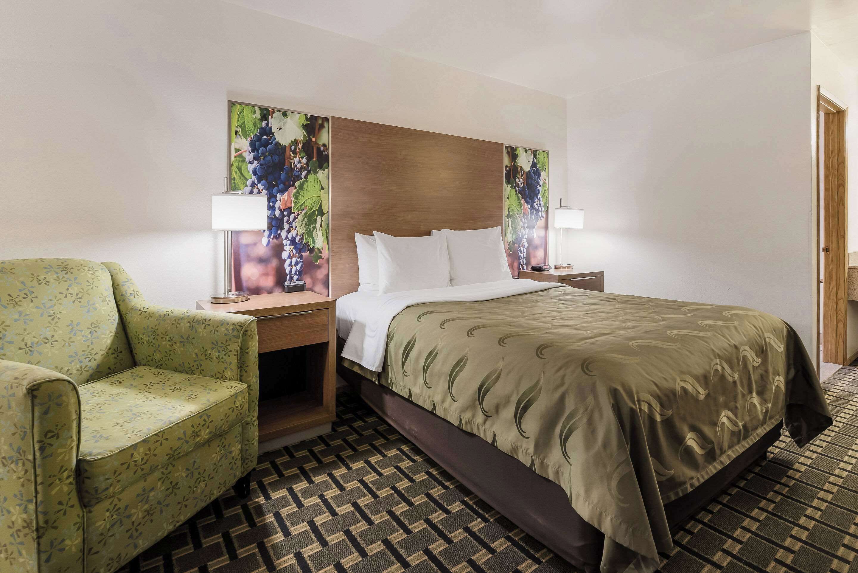 Quarto de hotel com uma cama, poltrona verde com padrão floral, cabeceira de madeira e duas luminárias fixadas na parede.