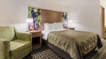 Quarto de hotel com uma cama, poltrona verde com padrão floral, cabeceira de madeira e duas luminárias fixadas na parede.
