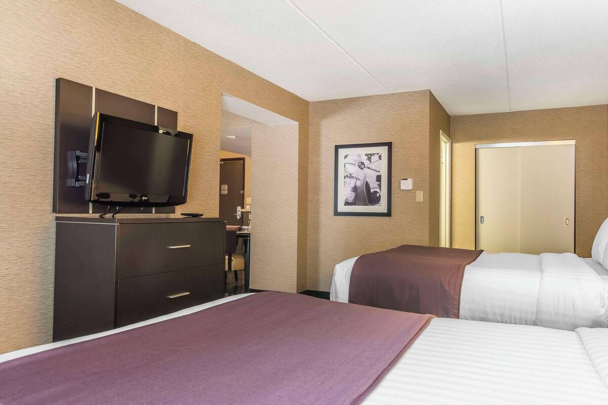 A hotel room with two beds, a TV on a dresser, a framed picture on the wall, and a door leading to another room.