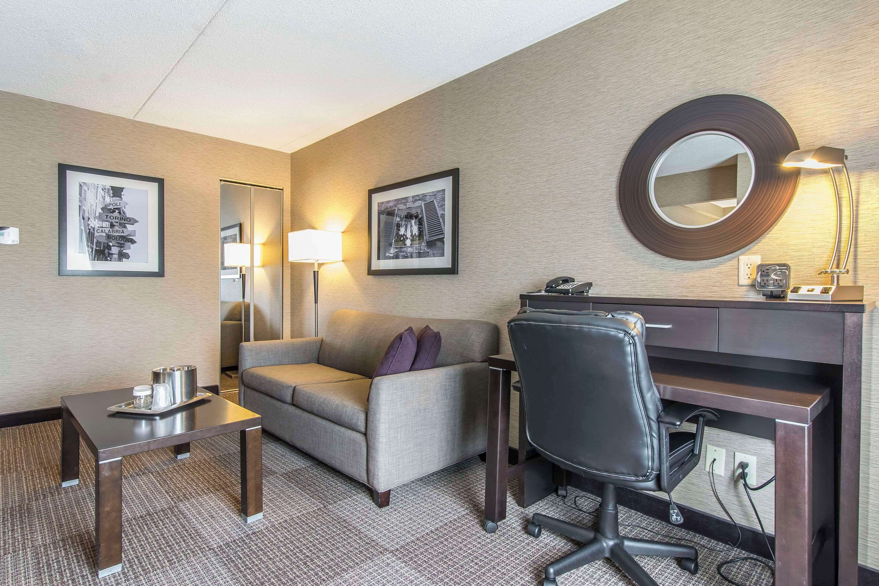 Ett hotellrum med ett skrivbord, en soffa, en stol, en lampa och inramade tavlor på väggarna.
