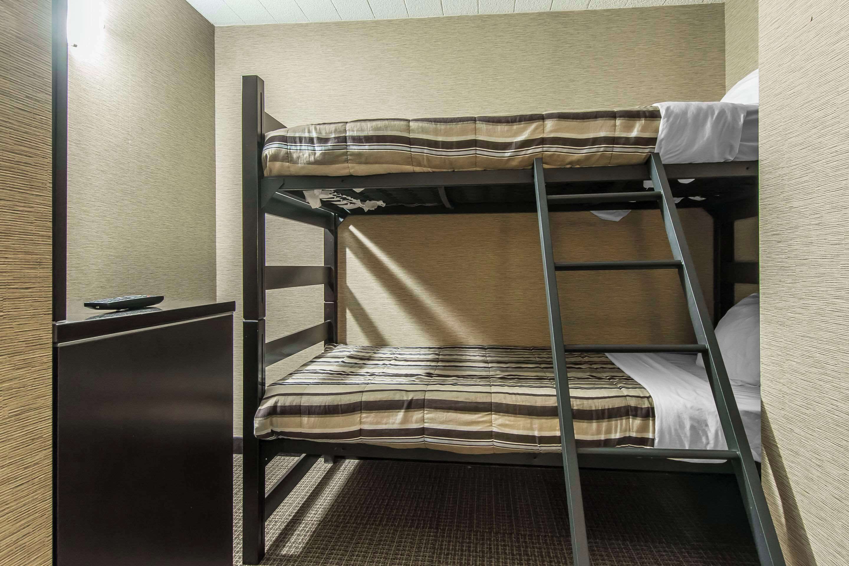 A bunk bed with a striped mattress and a ladder, a small desk with a remote control, and a carpeted floor.