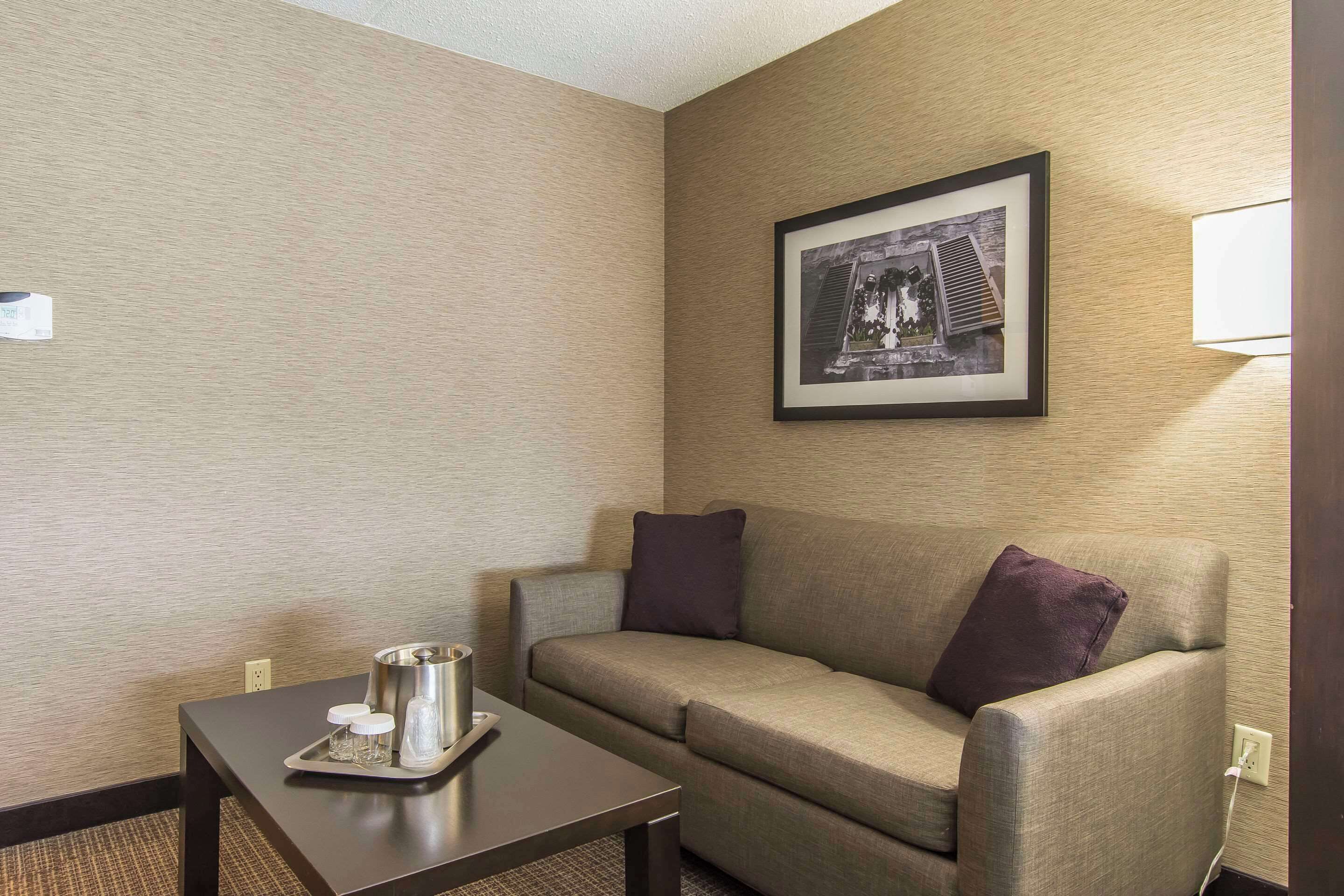A hotel room with a sofa, a small table with a tray of glasses and a pitcher, a framed picture on the wall, and a lamp.