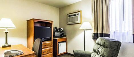 A hotel room with a wooden desk, a television, a microwave, a coffee maker, a chair, and a window with curtains.