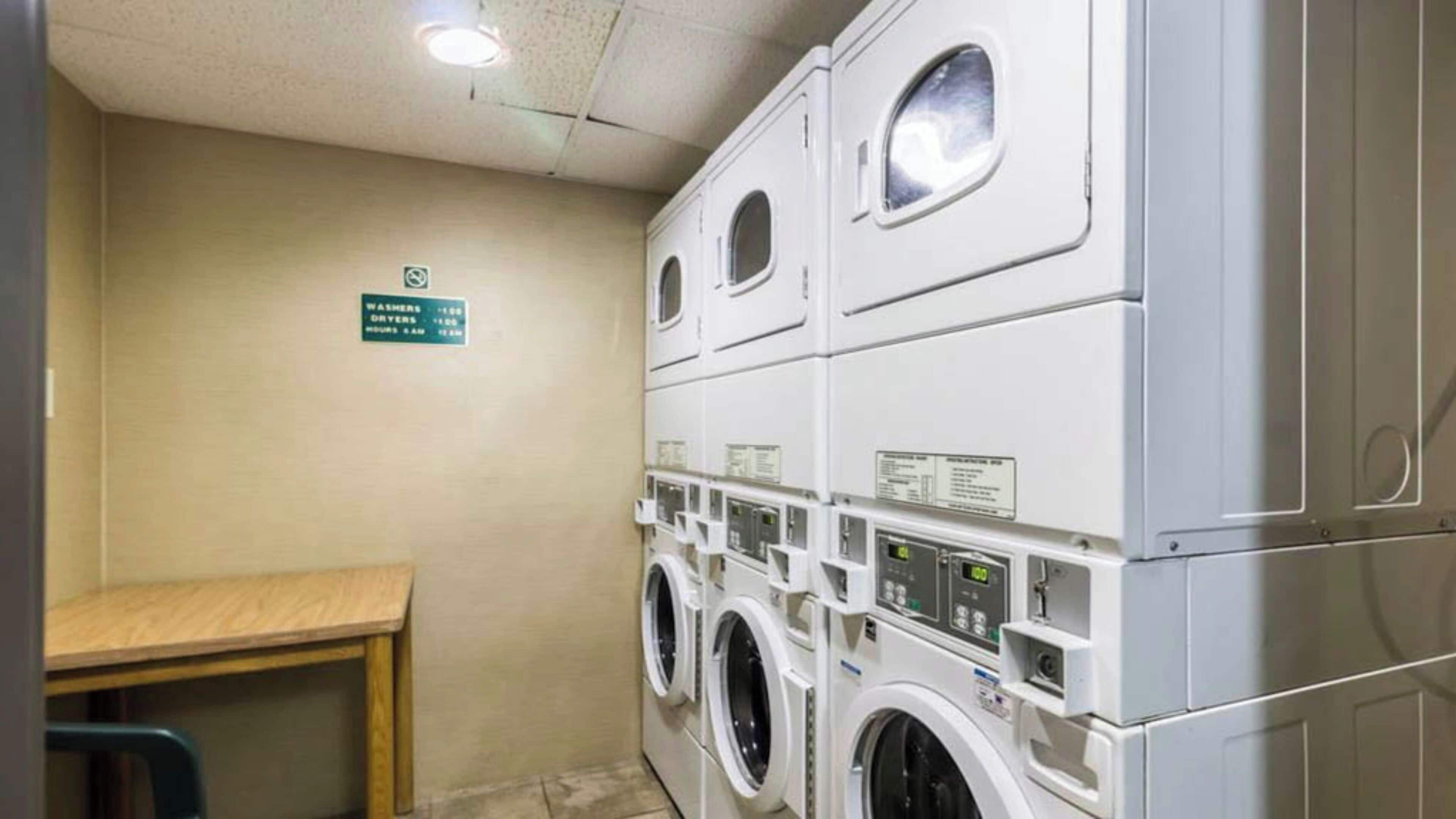 Un lavadero con varias lavadoras y secadoras apiladas, un banco de madera y un cartel que indica la cantidad de máquinas y sus capacidades.