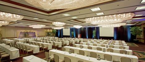 A banquet hall with rows of tables and chairs, large chandeliers, and a stage with curtains.
