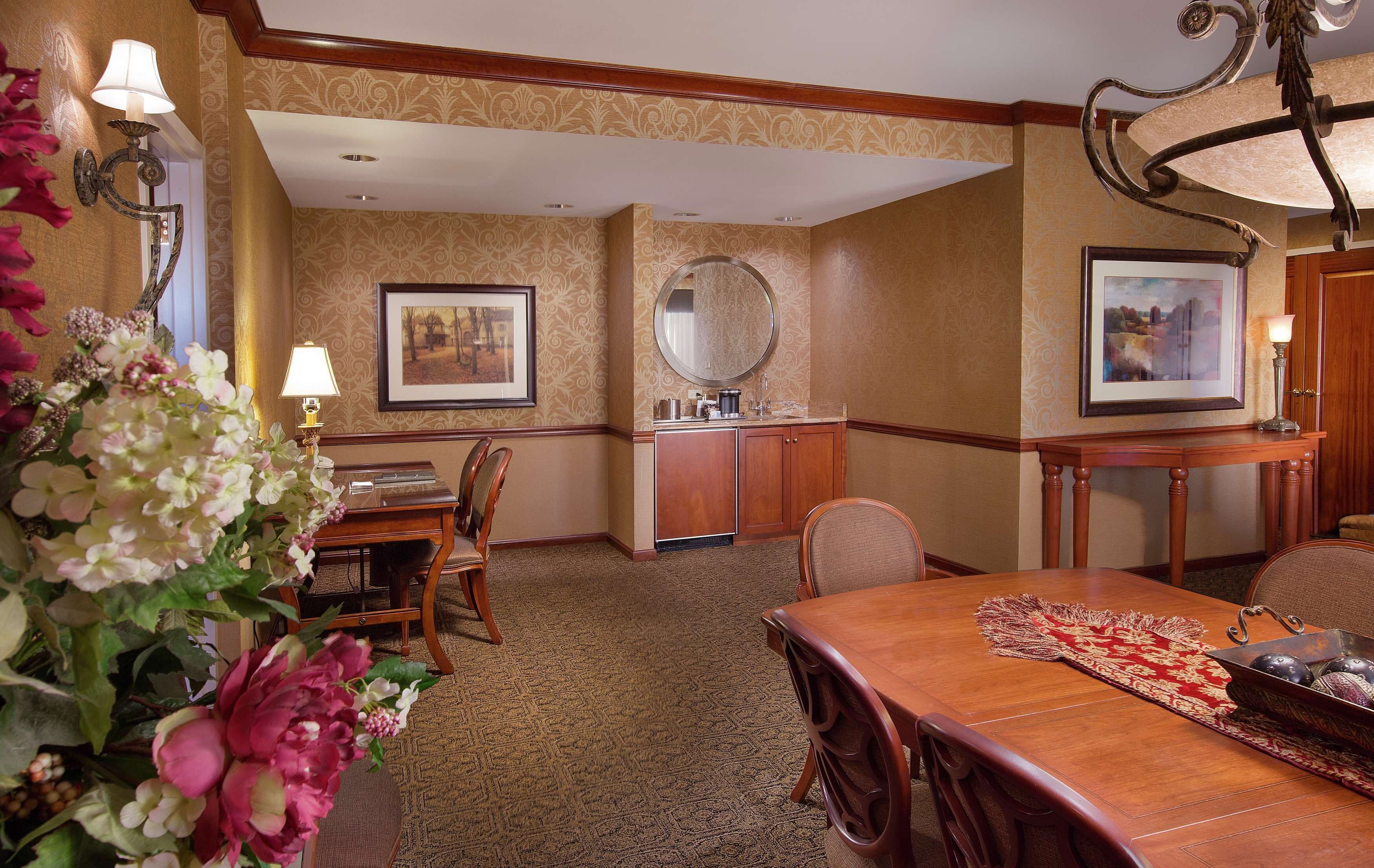 A hotel dining area with a wooden table, chairs, a vase of flowers, and wall-mounted lamps.