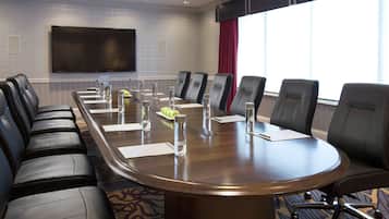 A conference room with a long table, multiple chairs, a flat-screen TV, and a large window with curtains.
