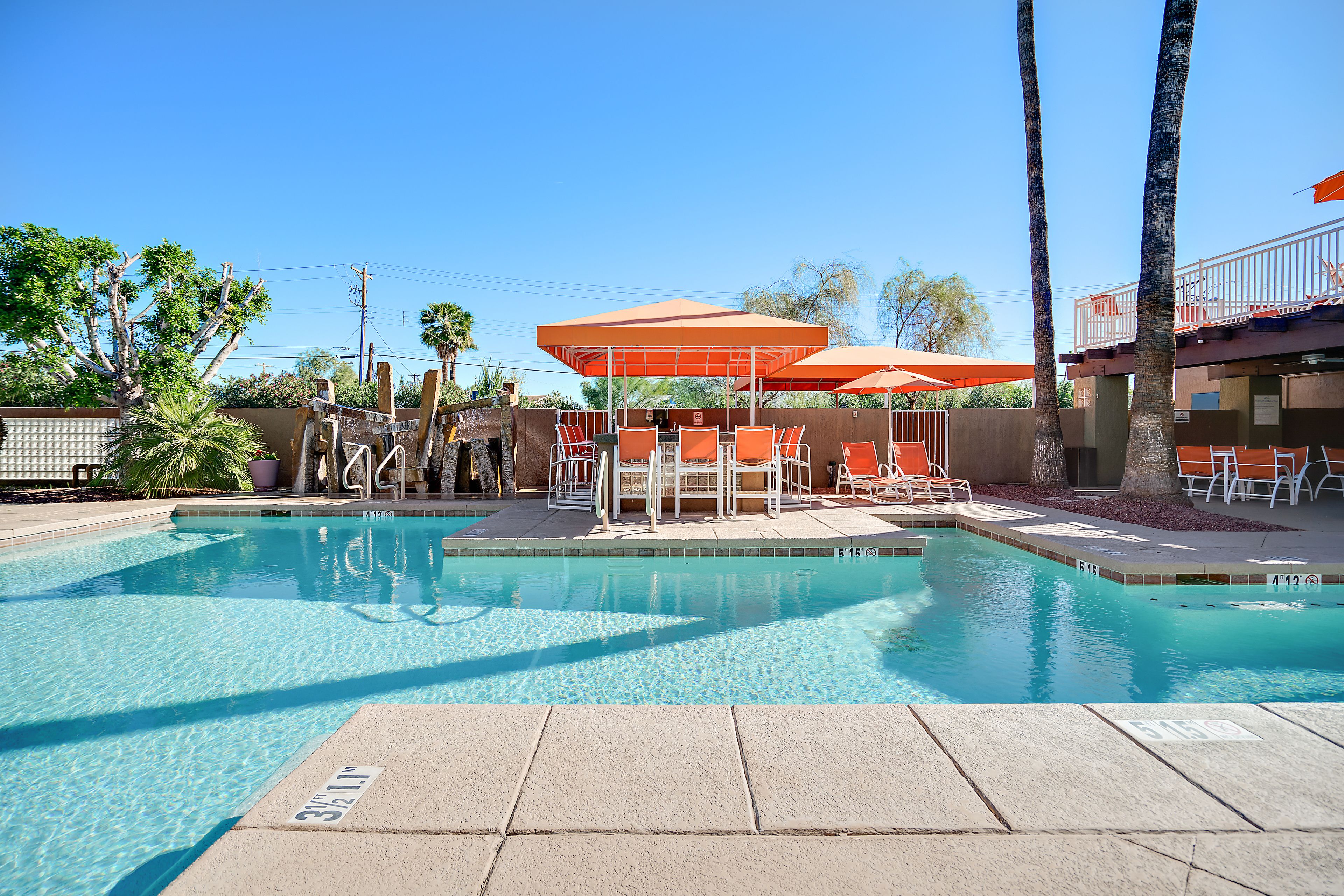 A clear swimming pool with lounge chairs and umbrellas, surrounded by palm trees and a building with a balcony.