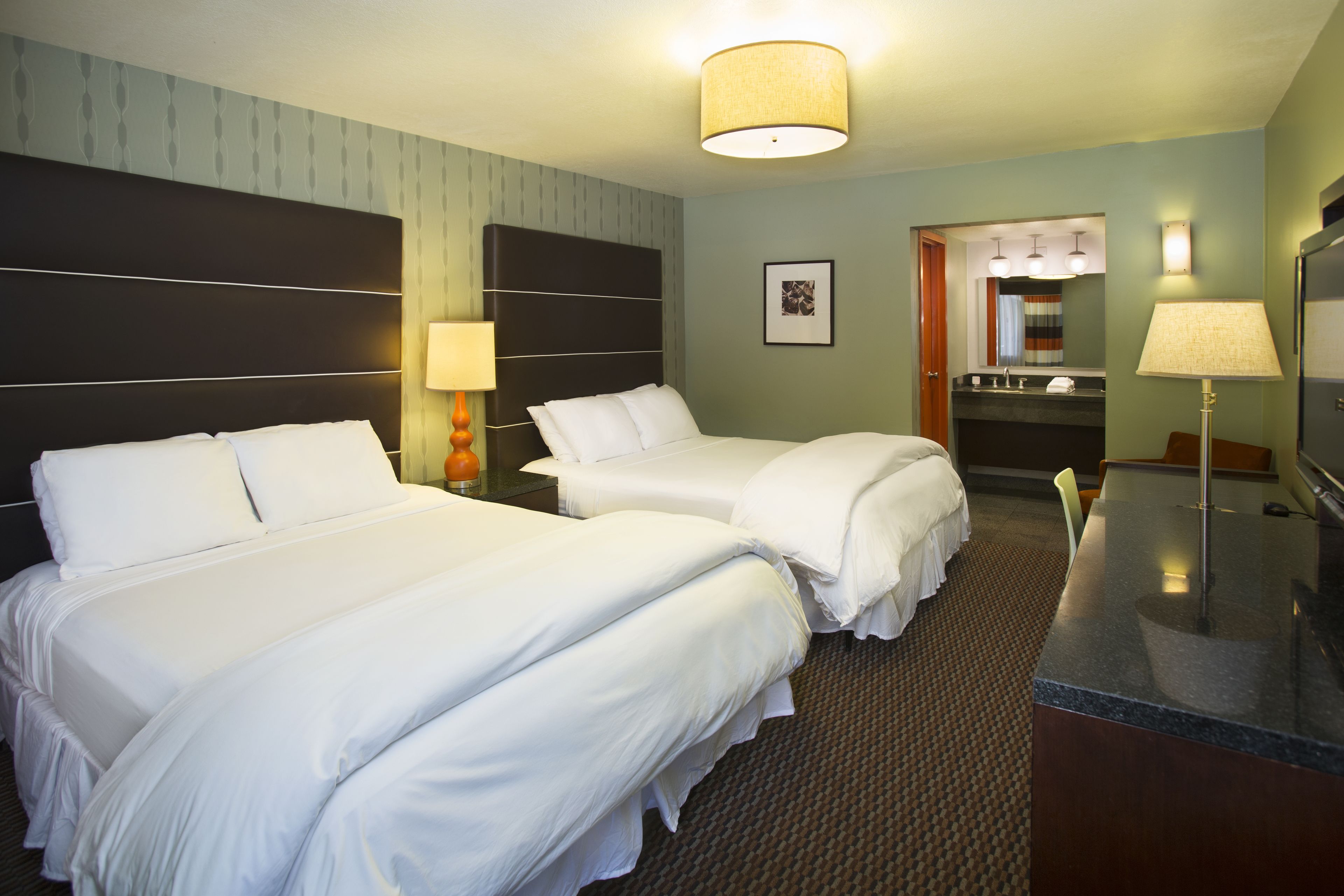 A hotel room with two beds, a desk, a chair, a lamp, a mirror, and a bathroom visible through an open door.