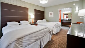 Habitación de hotel con dos camas, un escritorio, un espejo y un baño visible a través de una puerta abierta.