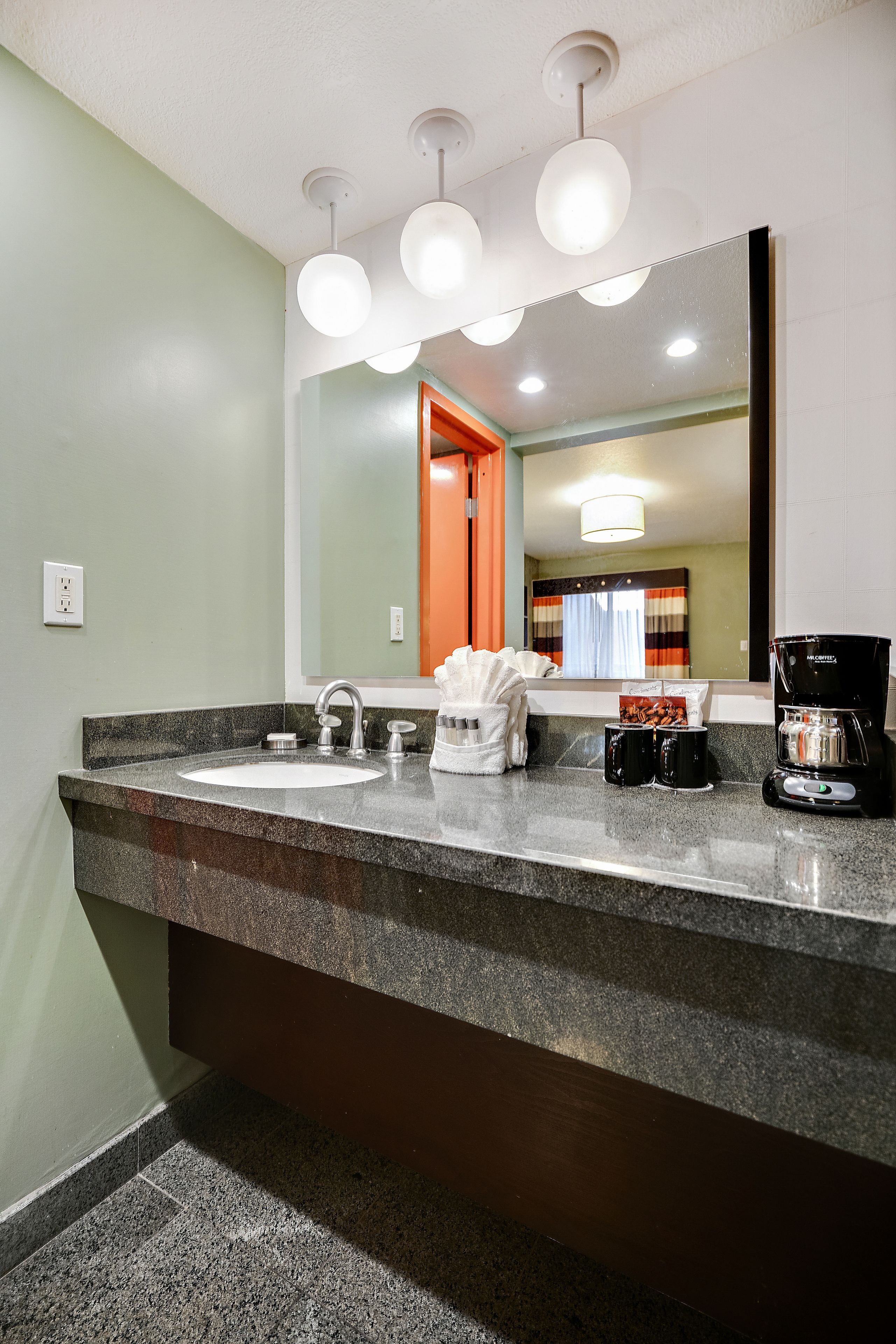 Un baño de hotel con encimera de granito, dos lavabos, cafetera y espejo que refleja una habitación con cama y cortinas.