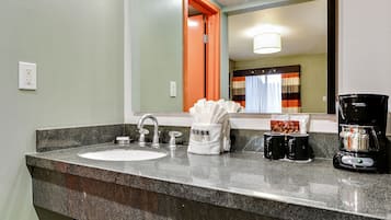 Un baño de hotel con encimera de granito, dos lavabos, cafetera y espejo que refleja una habitación con cama y cortinas.