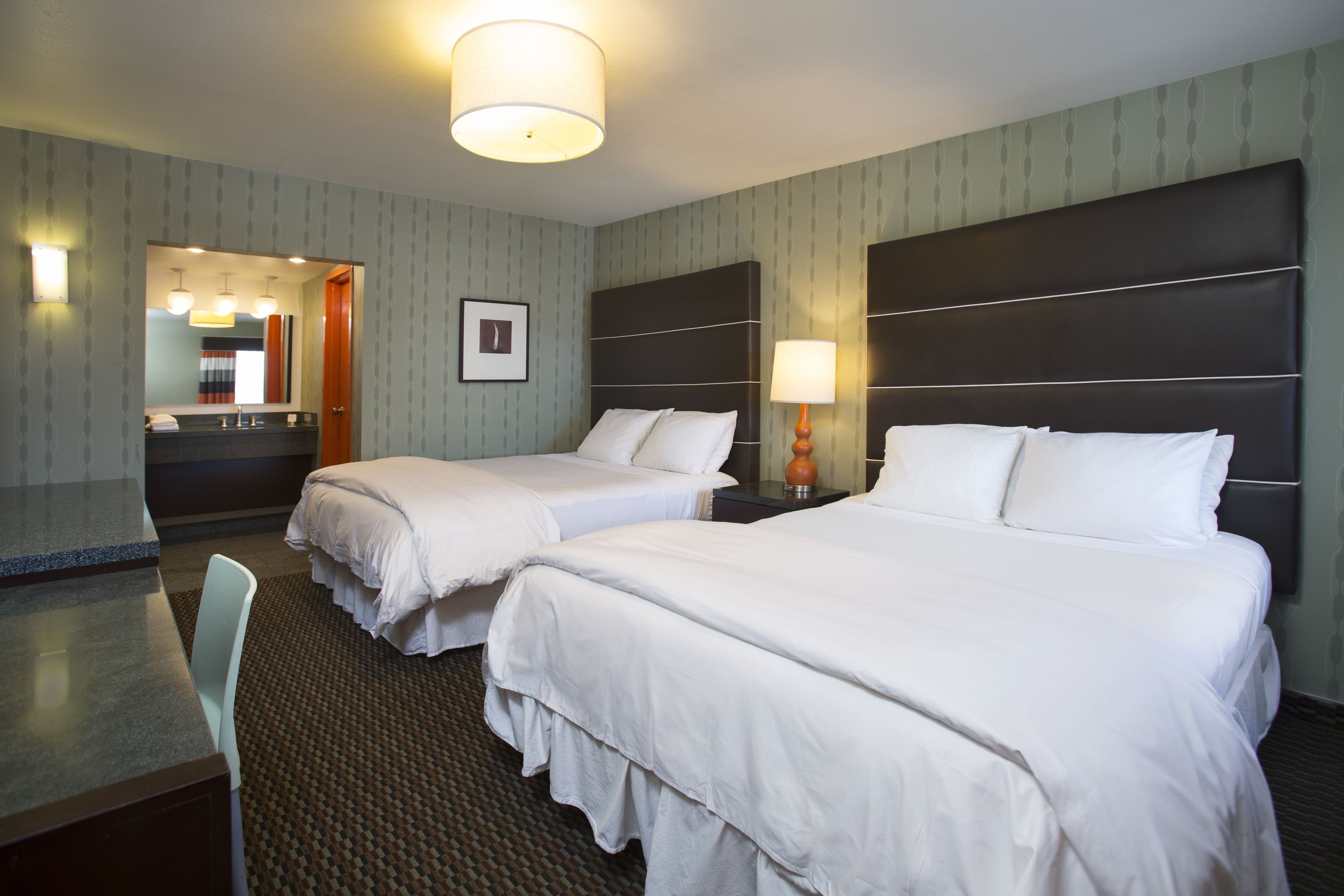 A hotel room with two beds, a desk, a mirror, and a bathroom visible through an open door.