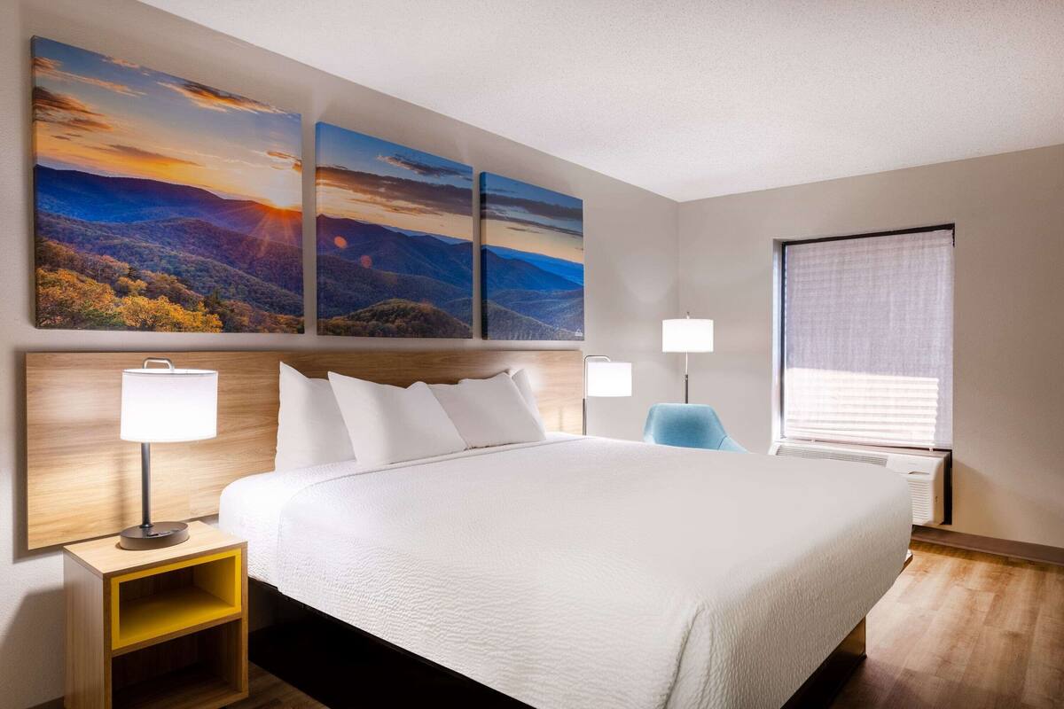 Ein Hotelzimmer mit einem großen Bett, zwei Nachttischen mit Lampen, einem Fenster mit Jalousien und drei Landschaftsbildern an der Wand.