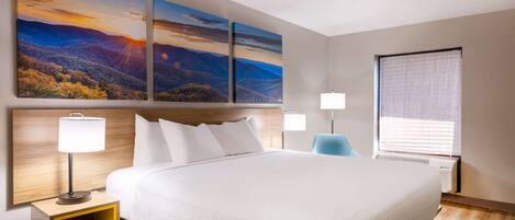 Habitación de hotel con una cama grande, dos mesitas de noche con lámparas, un tocador y una ventana con persianas, además de tres cuadros de paisajes en la pared.