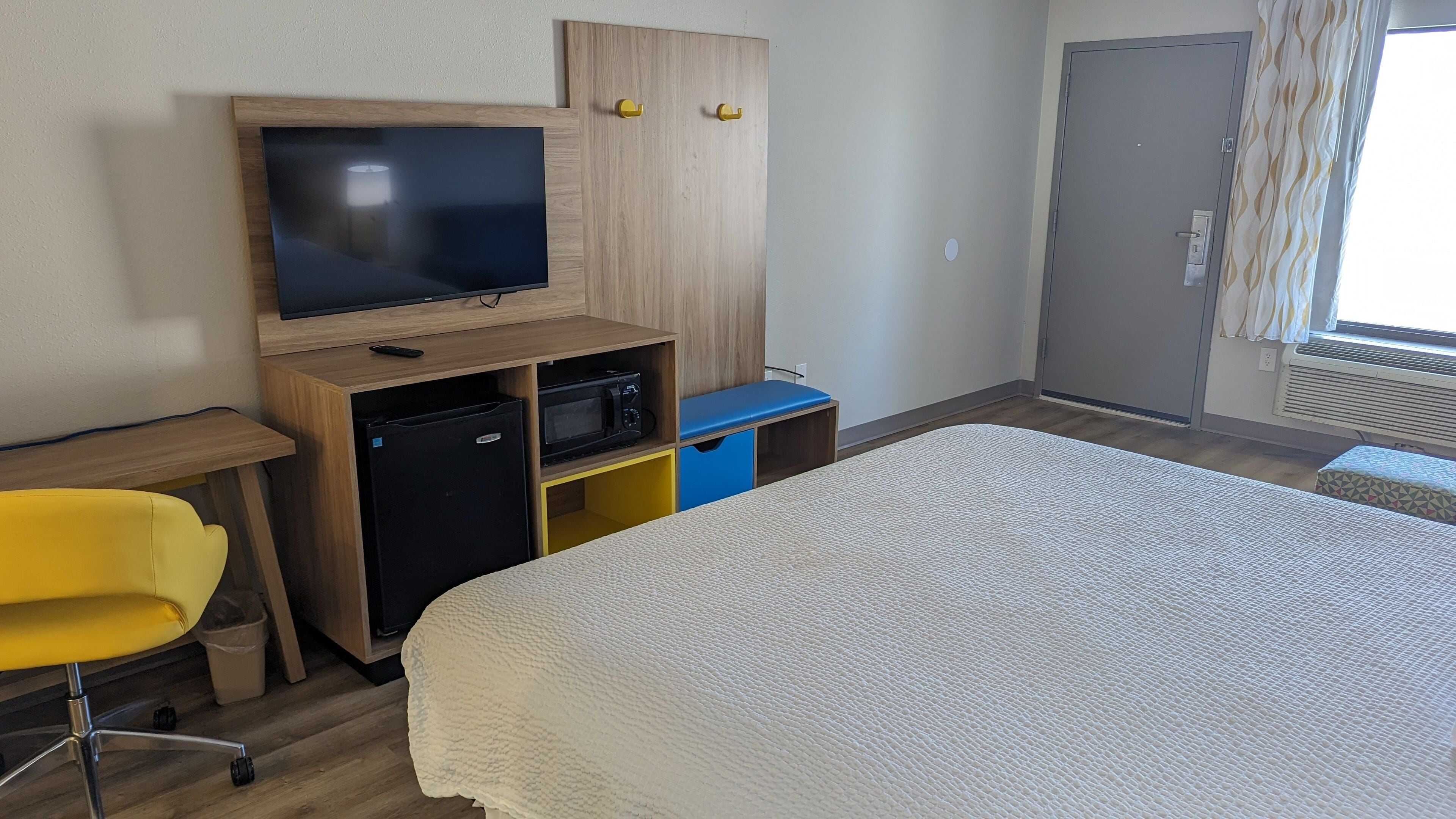 A hotel room with a bed, a desk with a television, a small refrigerator, and a yellow chair.