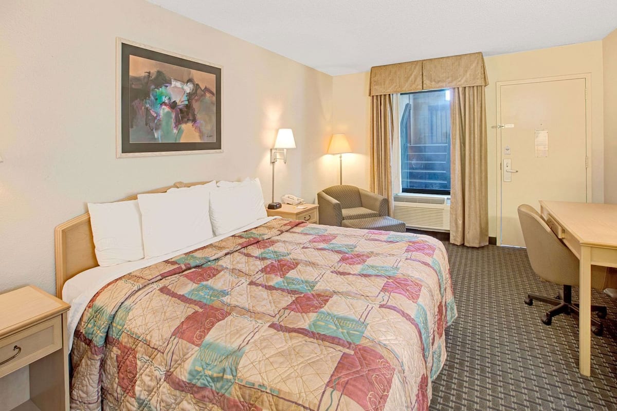 Ein Hotelzimmer mit Bett, Schreibtisch, Stuhl, Lampe, einem Gemälde und einem Fenster mit Vorhängen.