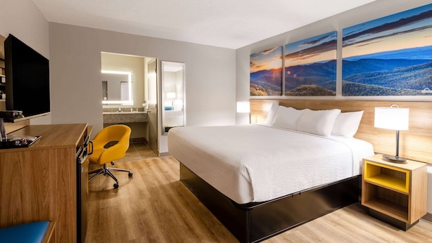 Une chambre d’hôtel avec un grand lit, un bureau avec une chaise, une télévision et un tableau représentant un paysage accroché au mur.