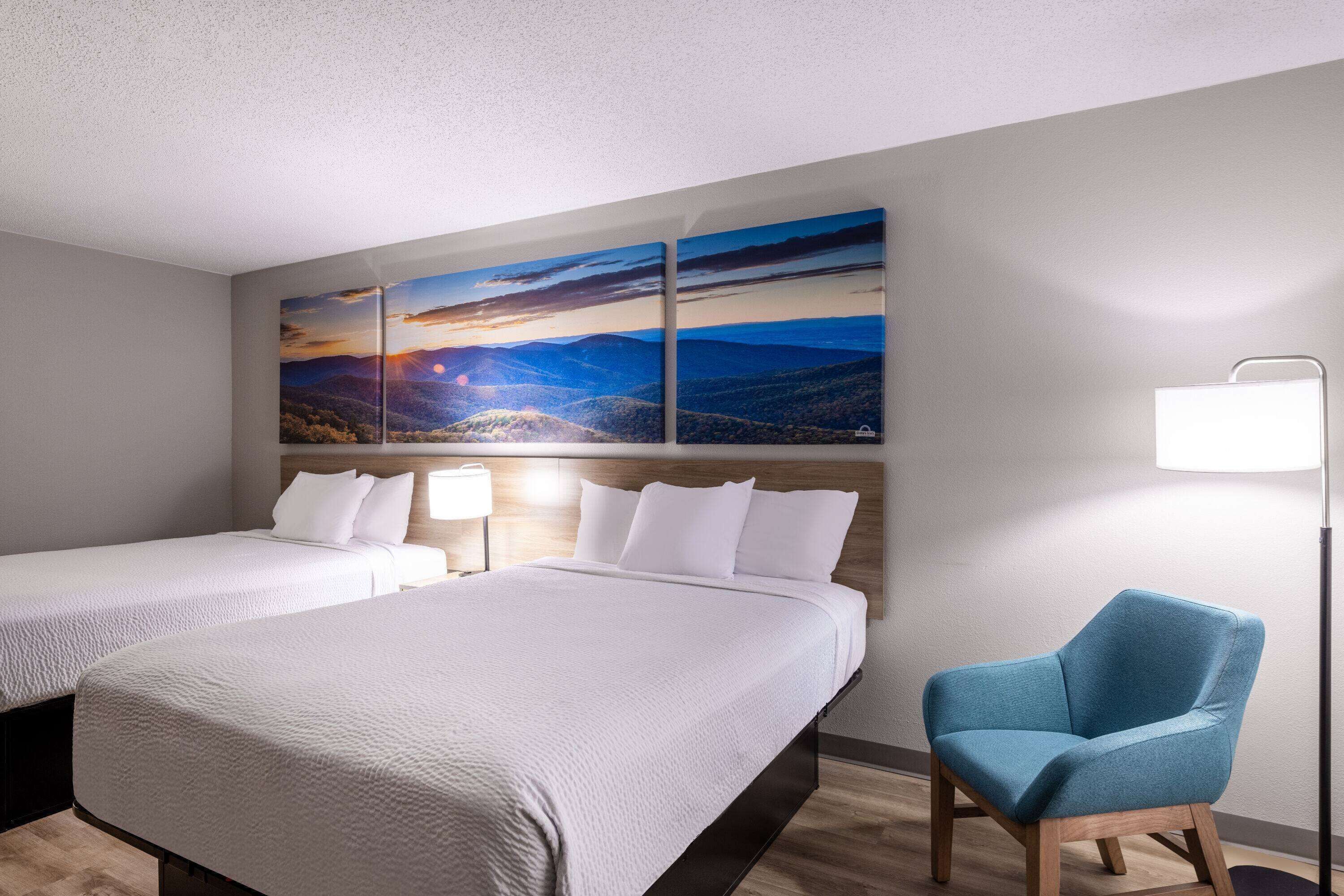 Une chambre d’hôtel avec deux lits, un fauteuil bleu et un grand tableau représentant un paysage accroché au mur.