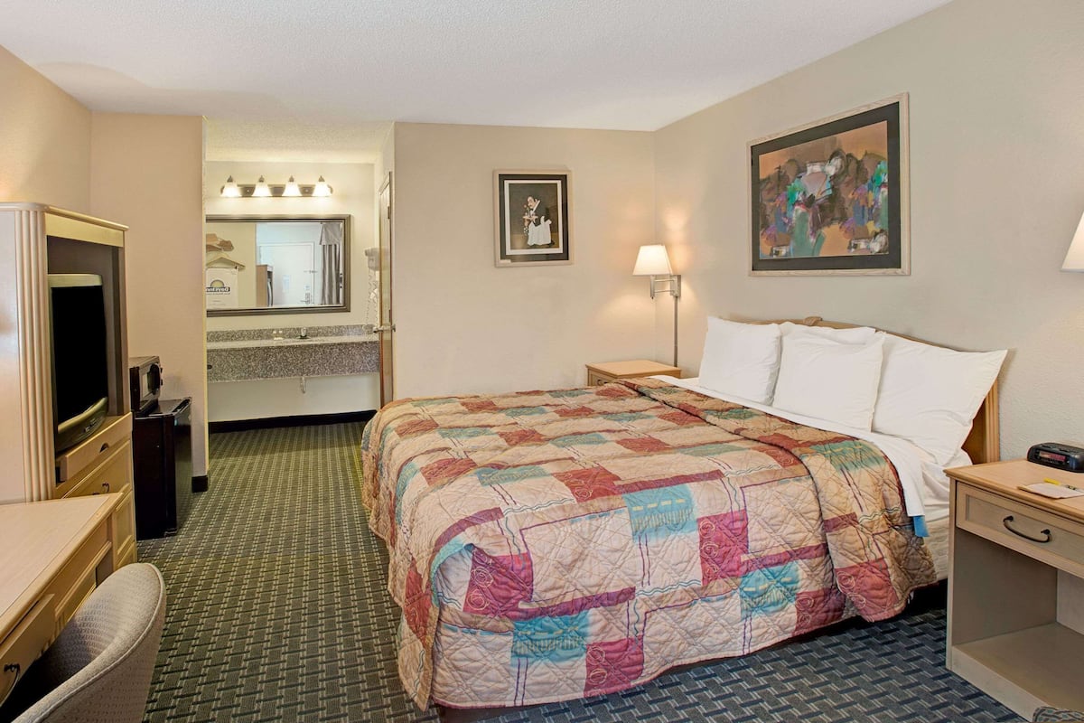Ein Hotelzimmer mit einem Bett, einem Schreibtisch, einem Stuhl, einer Kommode und einem Schminktisch mit Spiegel. An der Wand hängt ein Gemälde.
