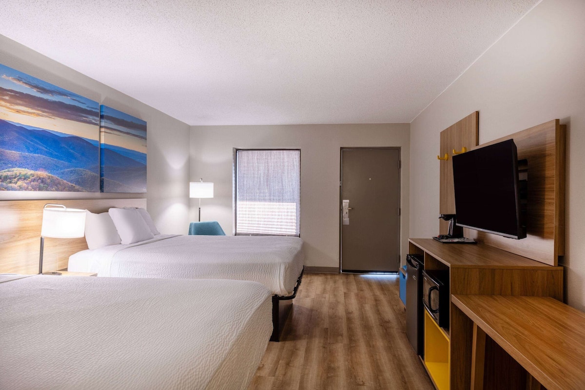 Ein Hotelzimmer mit zwei Betten, einem Fernseher, einem Schreibtisch, einer Lampe und einem Bild an der Wand.