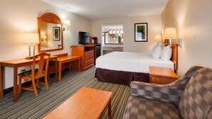 A hotel room with a bed, a desk, a chair, a sofa, a television, a mirror, and a bathroom visible through an archway.