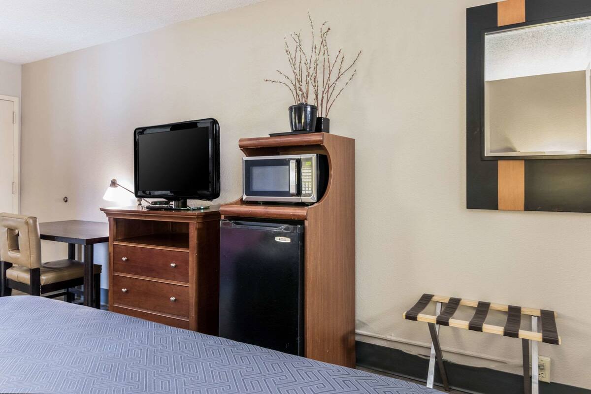 Quarto de hotel com cama, escrivaninha com televisão, micro-ondas, mini-geladeira, armário de madeira, cadeira e mesa de cabeceira.