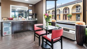 A modern hotel room with a kitchen area, a dining table with red chairs, and a large window offering a view of the street and buildings.