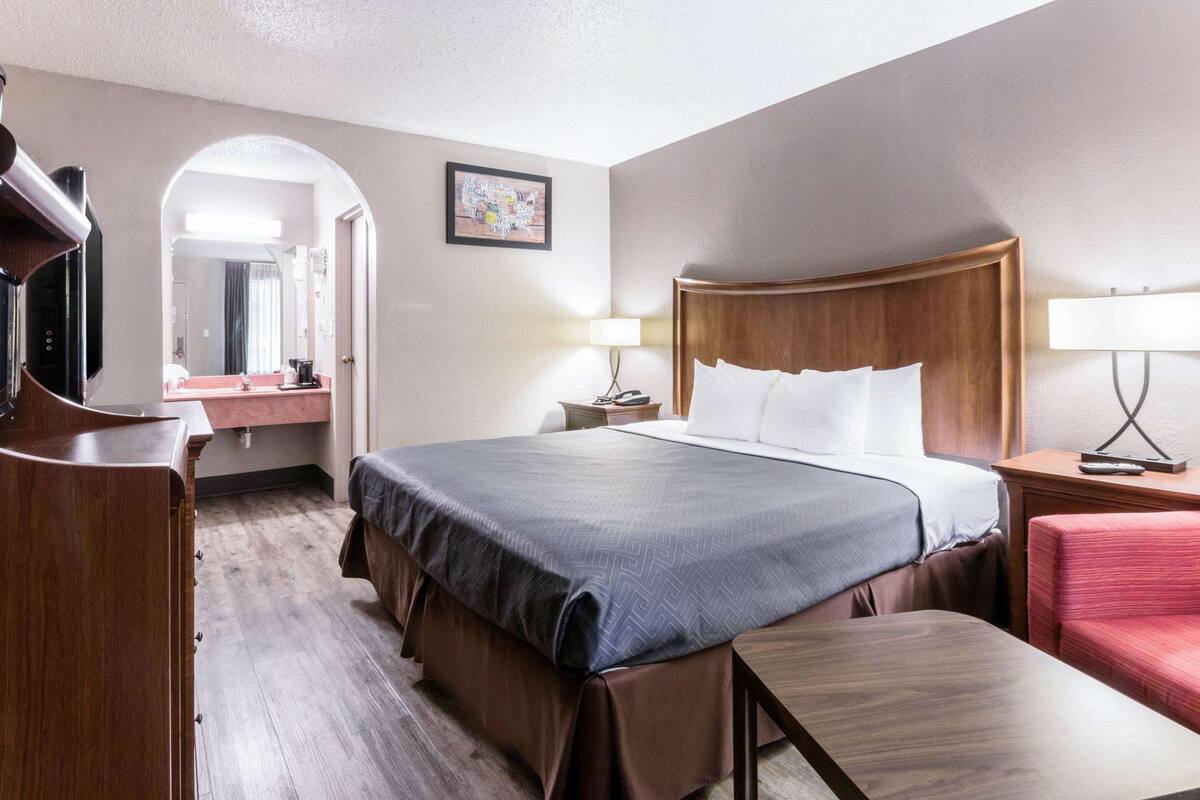 Quarto de hotel com uma cama grande, uma escrivaninha com cadeira, televisão, banheiro com espelho e um quadro na parede.