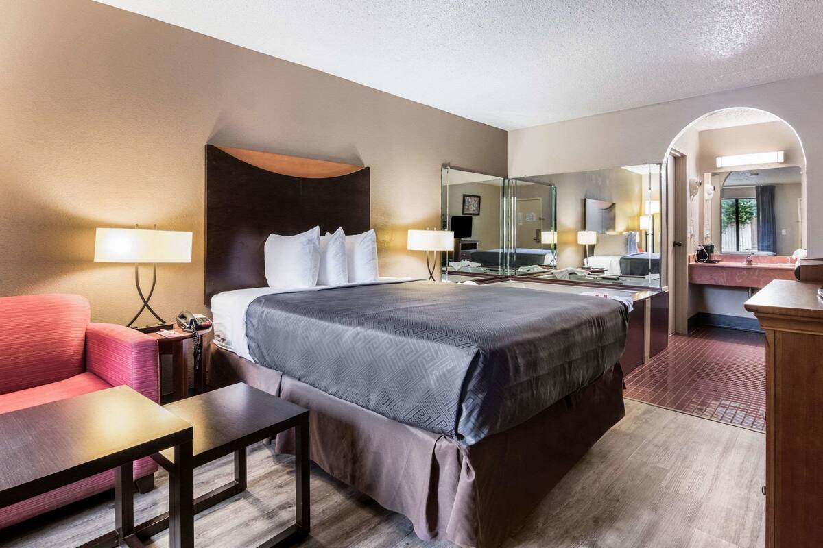 Quarto de hotel com uma cama grande, uma escrivaninha, uma cadeira, um sofá, um espelho e um banheiro visível por um arco.