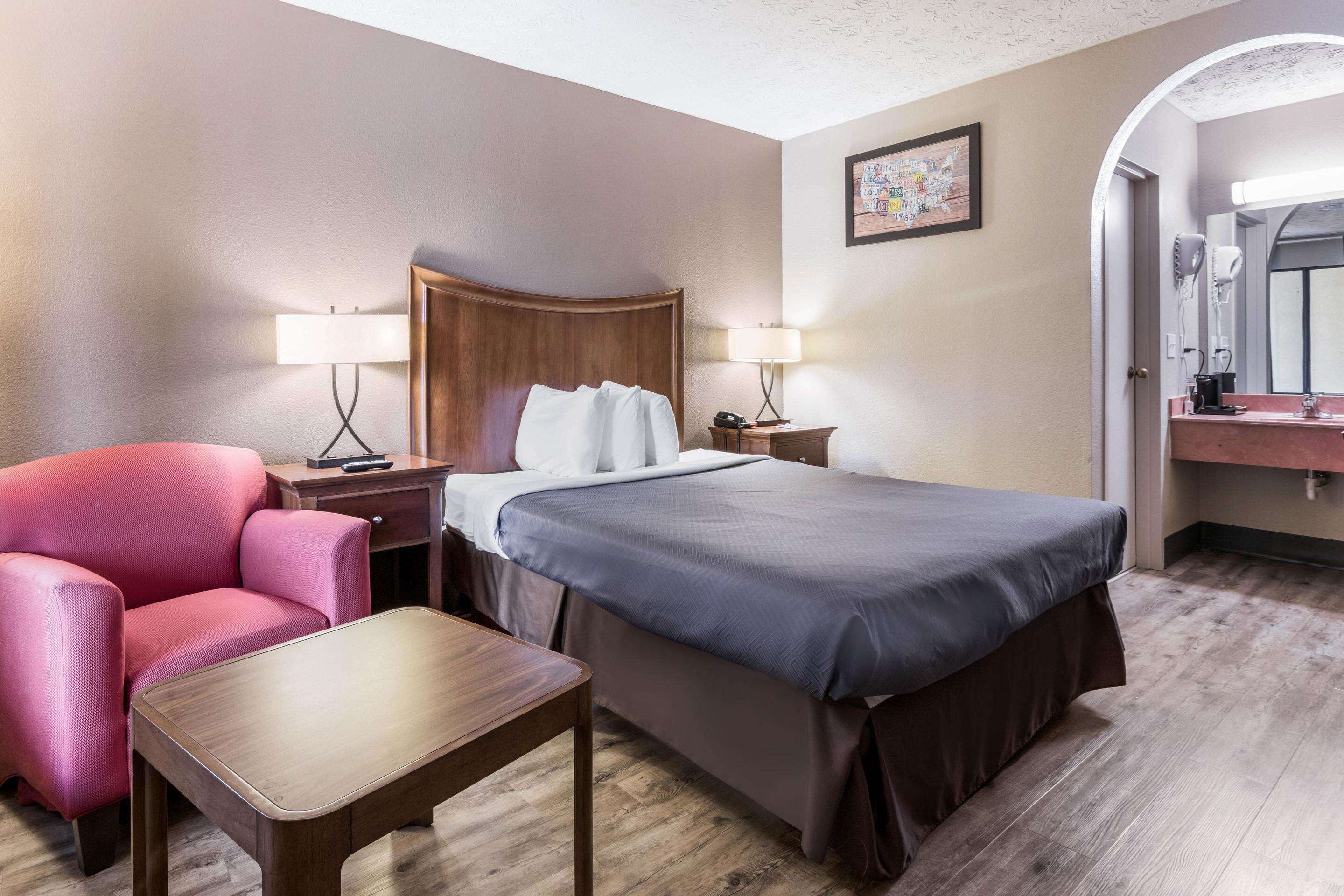 Habitación de hotel con cama, escritorio, silla, mesita de noche, lámpara, espejo y un cuadro en la pared.