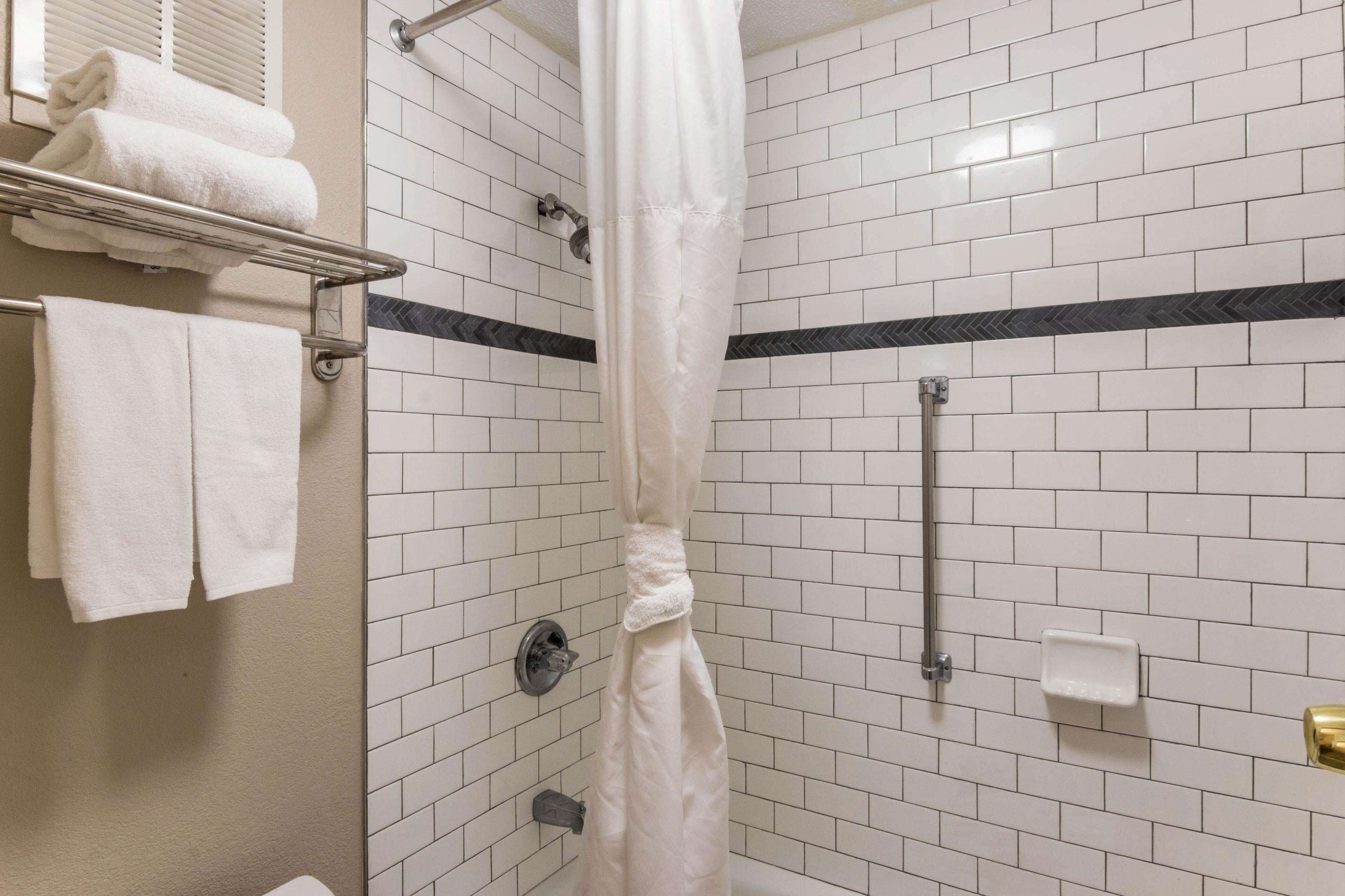 A shower area with white tiled walls, a shower curtain, a towel rack with folded towels, and a soap dispenser.