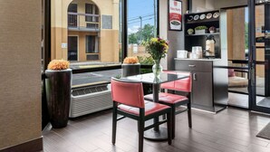 A modern dining area with a glass table and chairs, a large window with a view of a building, and a kitchen area with a coffee machine.