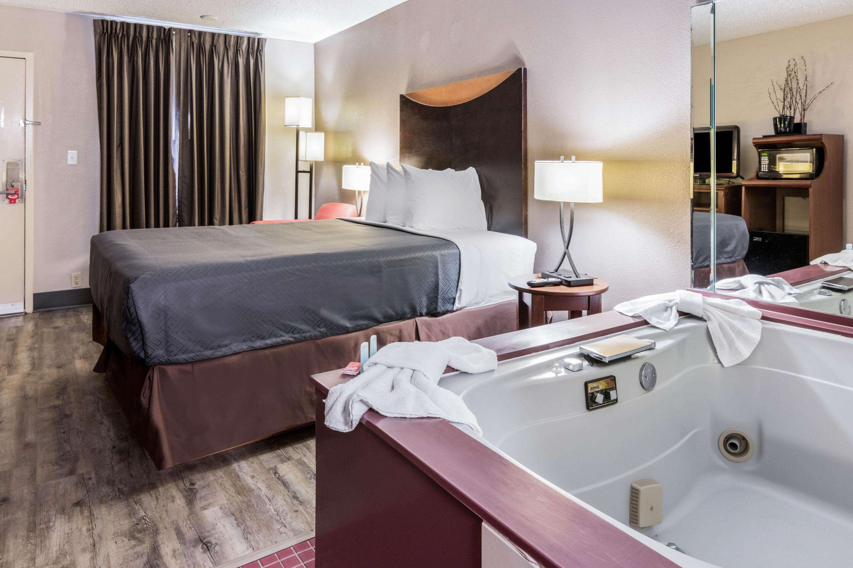 A hotel room with a large bed, a sitting area with a chair, a desk, a TV, a minibar, and a bathroom with a bathtub.