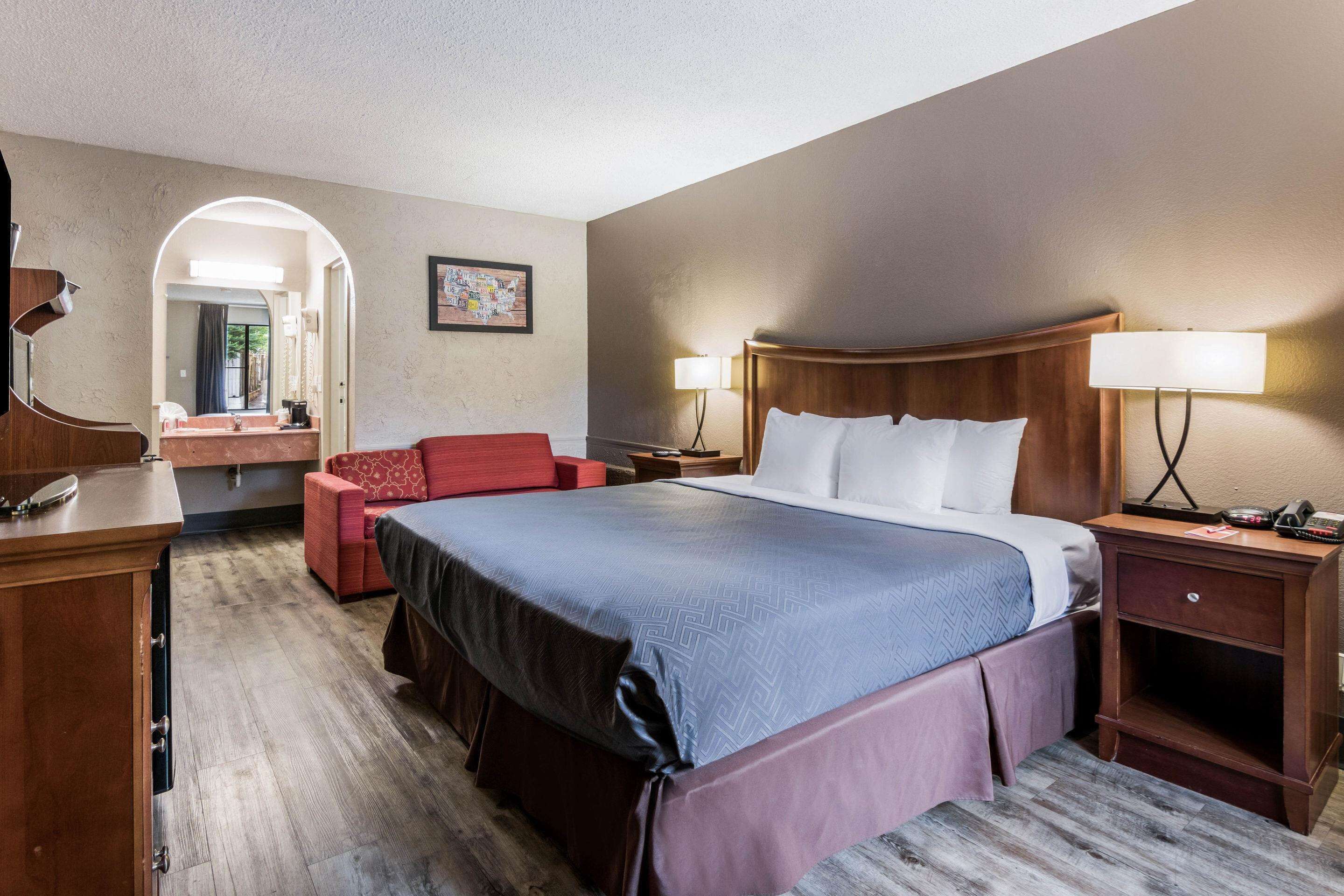 A hotel room with a large bed, a desk, a chair, a vanity with a mirror, and a picture on the wall.