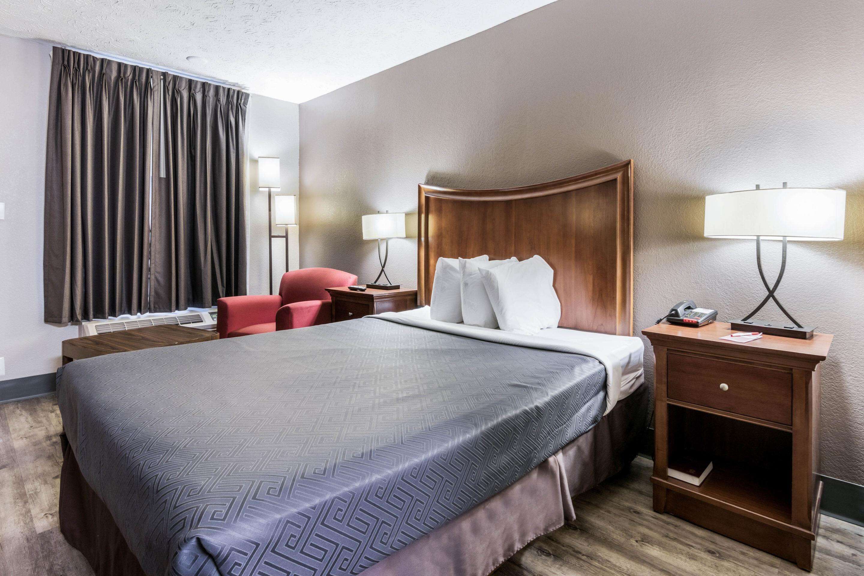 A hotel room with a large bed, a wooden nightstand, two bedside lamps, a red chair, and a desk with a lamp.
