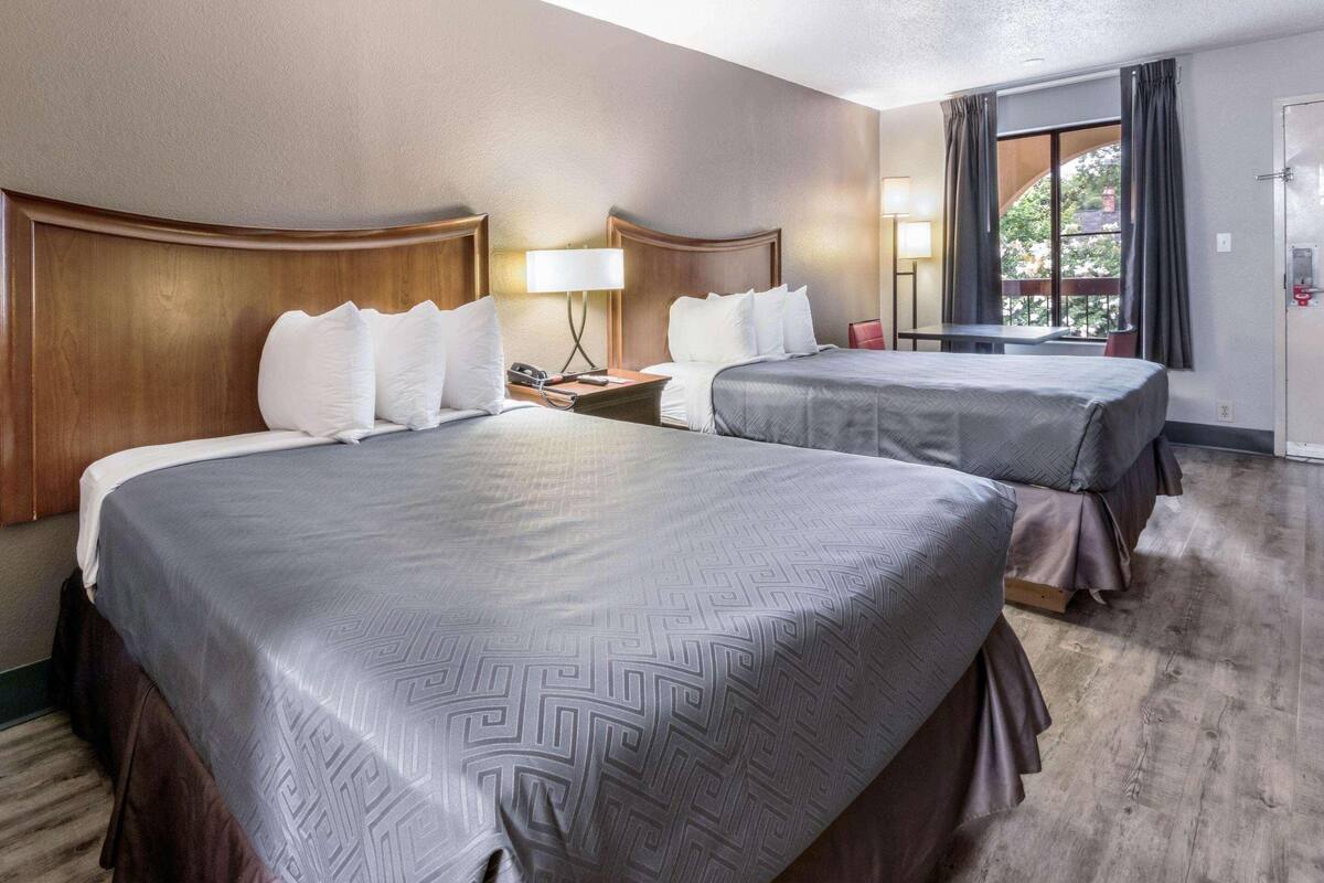 Quarto de hotel com duas camas, uma escrivaninha, uma cadeira, uma janela com cortinas e uma porta.